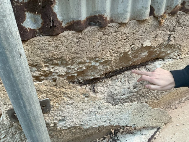 A person gestures towards a wall featuring a hole, with retaining walls visible in the background.